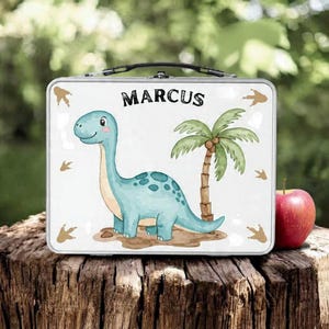 May include: A metal lunchbox with a cartoon dinosaur and palm tree design. The name "MARCUS" is printed at the top. The dinosaur is blue with green spots. Footprints and a red apple are also visible.