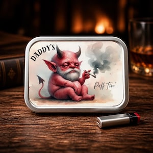 May include: A metal tin with the text "Daddy's Puff Tin" featuring a red devil illustration smoking a cigarette. A silver lighter is next to the tin on a wooden surface.