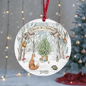 Pode incluir: Enfeite de Natal redondo com uma cena de floresta de inverno. Uma raposa, um esquilo e outros animais cercam uma árvore de Natal. O texto "Gracie's 1st Christmas" está em uma faixa, e o ano "2025" está na parte inferior. Uma fita vermelha está no topo.