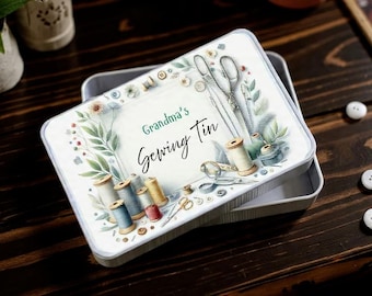 May include: A rectangular sewing tin with a floral and sewing-themed design. The lid features the text "Grandma's Sewing Tin" and illustrations of spools, scissors, and needles. The tin is open, revealing its interior.