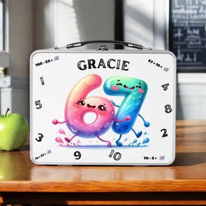 Könnte beinhalten: Eine weiße Metall-Lunchbox mit dem Namen "GRACIE" über den Zahlen "67" mit animierten Gesichtern. Die Zahlen sind rosa und blau mit Wasserspritzdetails. Zahlen 1-5, 8-10 und Matheaufgaben umgeben die Zahlen.