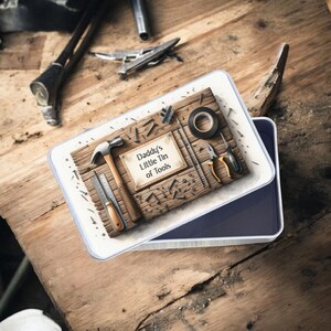 May include: A rectangular tin with a white border and a blue interior, open to reveal a wooden panel with tools. The panel features a hammer, tape, pliers, and a ruler. The text "Daddy's Little Tin of Tools" is printed on a label.