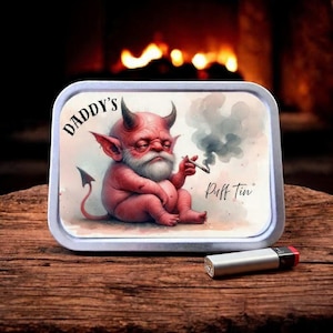 Puede incluir: Una lata de metal rectangular con una ilustración de un diablo de dibujos animados. El diablo rojo tiene cuernos, una barba blanca y está fumando un cigarrillo. La lata dice "DADDY'S" y "Puff Tin". Un encendedor plateado con tapa roja está al lado.