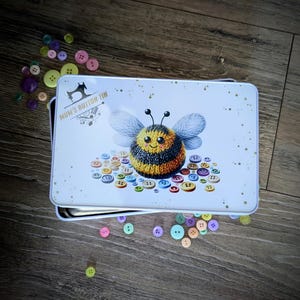 Puede incluir: Una lata rectangular blanca con una ilustración de abeja y el texto "MUM'S BUTTON TIN". La lata está rodeada de botones coloridos de varios tamaños. La abeja es amarilla, negra y azul.