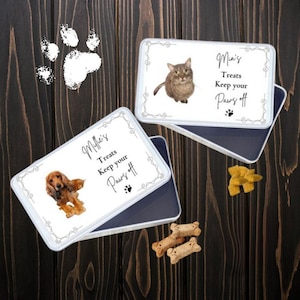 Op de afbeelding: Twee rechthoekige blikken voor dierlijke lekkernijen met deksels, één met een bruine hond en de andere met een grijze kat. Beide blikken hebben de tekst "Millie's Treats Keep your Paws off" en een pootafdruk.