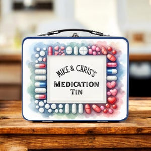 Puede incluir: Una caja de medicamentos blanca y azul con un asa negra, con el texto "MIKE & CHRIS'S MEDICATION TIN". La caja está decorada con pastillas de estilo acuarela en varios colores, incluyendo rojo, rosa, blanco y azul.