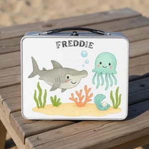 Op de afbeelding: Een zilveren metalen lunchbox met een handvat, met een cartoon haai, kwal en zeegroenten. De naam "FREDDIE" staat in zwarte letters bovenaan. Het ontwerp staat tegen een witte achtergrond.