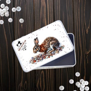 May include: A white rectangular tin featuring a watercolour illustration of a brown rabbit and assorted buttons. The tin is labelled "Nan's Button Tin" with a sewing machine and musical notes. White buttons are scattered on a dark wooden surface.