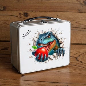 Op de afbeelding: Een zilveren metalen lunchbox met een zwarte handgreep. De voorkant heeft een kleurrijke illustratie van een dinosaurus die door een muur breekt en een rode appel vasthoudt. De naam "Mark" staat linksboven gedrukt.