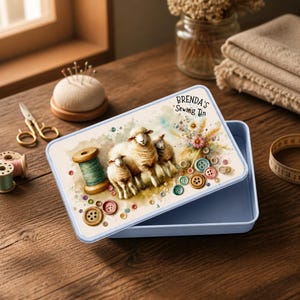 May include: A light blue sewing tin with a watercolor illustration of sheep, a spool of green thread, and colorful buttons. The tin is labeled "Brenda's Sewing Tin". Other sewing supplies are in the background.