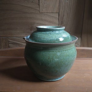 May include: A handcrafted, green ceramic jar with a matching lid. The jar has a rounded body and a wide, flat rim. The lid sits snugly on top. The piece is displayed on a wooden surface against a wooden backdrop.