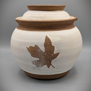Peut inclure: Un pot en céramique fait à la main avec un couvercle. Le pot est blanc avec des accents bruns et un motif de feuille marron. Le pot mesure environ 18 cm de haut et 15 cm de large.