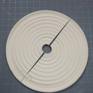 May include: A light grey ceramic tree collar, split in two pieces, with a central hole and concentric circular ridges. The collar has embossed leaf designs on each side. The collar is on a blue cutting mat.