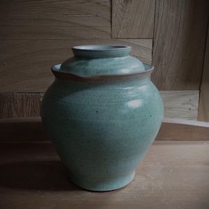 Può includere: Un vaso in ceramica verde acqua fatto a mano con coperchio. Il vaso ha un corpo arrotondato e un bordo leggermente svasato, con una fascia marrone a contrasto sulla parte superiore. Il coperchio si adatta perfettamente. Il vaso è su uno sfondo di legno.