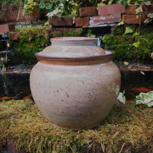 May include: A round ceramic jar with a lid, featuring a glossy beige glaze with brown speckles and a dark brown rim. The jar has a wide, bulbous body and a flat, circular lid that sits securely within the opening of the container.