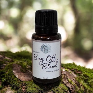 May include: A brown glass bottle with a black cap, labeled "Bug Off Blend Essential Oil". The label includes the text "100% Pure" and "15 ml". The bottle sits on a moss-covered log against a blurred green background.