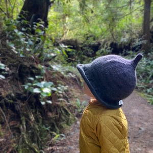 May include: A child wearing a dark gray, pointed hat and a yellow quilted jacket stands in a forest setting. The child is looking up and to the right.