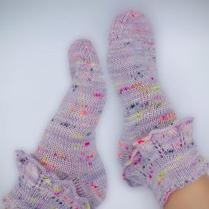 May include: A pair of lavender knitted socks with colourful flecks. The socks have a ruffled cuff detail. The socks are made of a soft, textured yarn and are displayed against a white background.