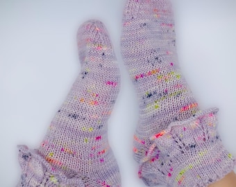May include: A pair of lavender-colored knitted socks with colorful flecks. The socks have a ruffled cuff detail. The socks are made of a soft, textured yarn and are displayed against a white background.