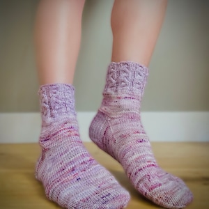 Può includere: Un paio di calzini a righe viola e bianche con un motivo a treccia in cima. I calzini sono indossati ai piedi di una persona.