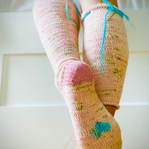 May include: A pair of pink and white knee-high socks with blue ribbon ties. The socks are knit with a speckled yarn and have a colorful pattern.