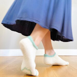 May include: A pair of white ankle socks with a light blue trim. The socks are worn by a person wearing a blue skirt.