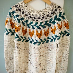 May include: A white knitted jumper with a colourful pattern featuring fox faces and a green and blue geometric design.