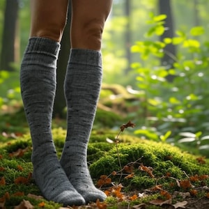 May include: A pair of gray knee-high socks. The socks are made of a thick, textured material and are pulled up to the knees. The socks are worn outdoors on a bed of green moss and fallen leaves, with a blurred forest background.