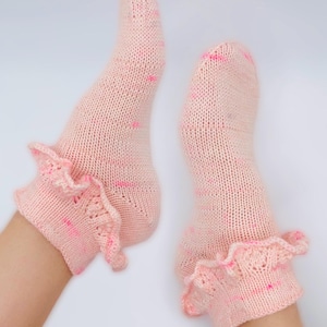 May include: A pair of pale pink knitted socks with a frilled cuff. The socks feature a speckled pattern of darker pink dots and dashes. The socks are shown against a white background.