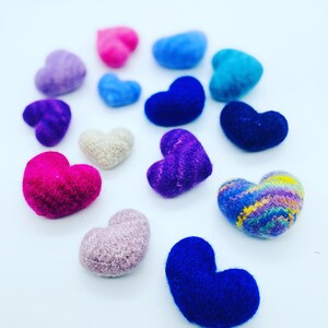 May include: A collection of felt hearts in various colors, including pink, purple, blue, and white. The hearts are arranged in a semi-circle on a white background.