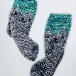 May include: A pair of gray and teal knitted socks with a cat face design. The socks are made with a soft, textured yarn.
