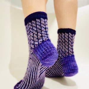 Può includere: Un paio di calzini lavorati a mano in tonalità viola e lavanda. I calzini presentano un motivo a coste sul piede e un design strutturato sulla gamba, con un bordo blu scuro. I calzini sono esposti su uno sfondo bianco.