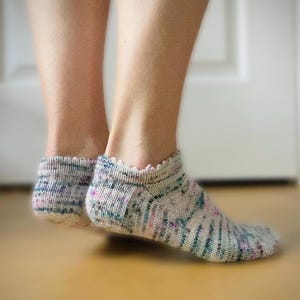 May include: A pair of ankle socks with a hand-knitted appearance. The socks feature a speckled pattern with shades of gray, pink, and teal. The top edge has a scalloped design. The socks are on a wooden surface.