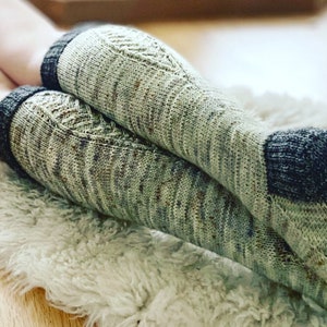 May include: A pair of hand-knitted socks featuring a variegated light green yarn with subtle brown and blue speckles. The socks have a dark charcoal grey cuff and heel, with a textured herringbone stitch pattern running along the top of the foot.