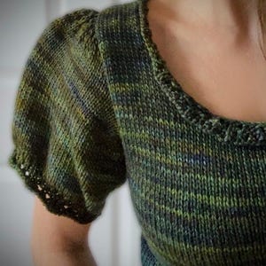 May include: A green knit top with short, puffed sleeves. The top features a square neckline with a scalloped edge. The yarn has a variegated pattern of green, blue, and yellow hues, creating a textured appearance. The sleeves have a delicate, openwork trim.