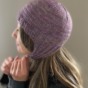 Może przedstawiać: Czapka z dzianiny w kolorze fioletowym i białym z długim nausznikiem, który można opuścić, aby zakryć uszy i szyję.