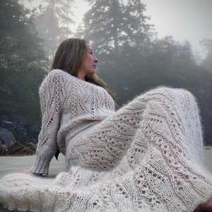Könnte beinhalten: Ein cremefarbenes Strickset aus Pullover und Rock. Der langärmlige Pullover hat einen Rundhalsausschnitt und aufwendige Lochmuster. Der passende Rock reicht bis zum Boden und weist ebenfalls ein detailliertes Lochmuster auf. Das Set wird an einem Strand mit nebligem Waldhintergrund getragen.