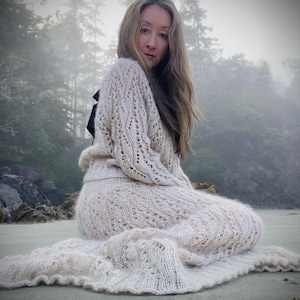 Puede incluir: Un conjunto de suéter y falda de punto color crema con un diseño calado. El conjunto se usa en una playa arenosa con un fondo de bosque brumoso. La falda tiene forma de sirena y el suéter tiene mangas largas.