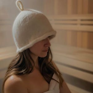 May include: A white sauna hat with a rounded crown and a small loop at the top. The hat is made of a soft, textured material. The person is in a sauna, wrapped in a white towel.