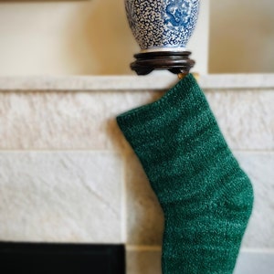 Op de afbeelding: Een groene kerstsok hangt aan een witte schouw. De sok is gemaakt van een pluche materiaal en heeft een licht getextureerd oppervlak.