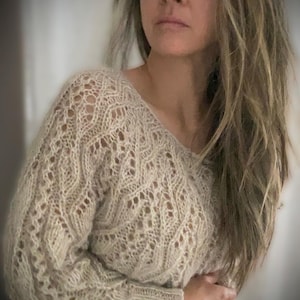 Könnte beinhalten: Ein beigefarbener Pullover mit offenem Gewebe und V-Ausschnitt. Der Pullover hat ein strukturiertes, gemustertes Design und lange Ärmel. Das Strickwarenmaterial wirkt weich und bequem, geeignet für kühleres Wetter. Der Gesamtstil ist lässig und entspannt.