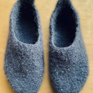 May include: A pair of dark gray felted wool slippers with a textured, fuzzy surface. The slippers feature a rounded toe design, a deep opening for the foot, and a slightly raised heel tab at the back, showcasing a dense, uniform material construction.