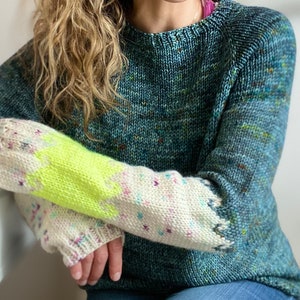 Puede incluir: Un suéter tejido a mano en tonos turquesa y azul con detalles moteados. Las mangas presentan un patrón color crema con una sección amarilla neón brillante. Una prenda acogedora hecha a mano.