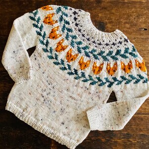 May include: A white knitted jumper with a colourful Fair Isle pattern featuring orange, green and blue accents. The jumper has a crew neck and long sleeves.