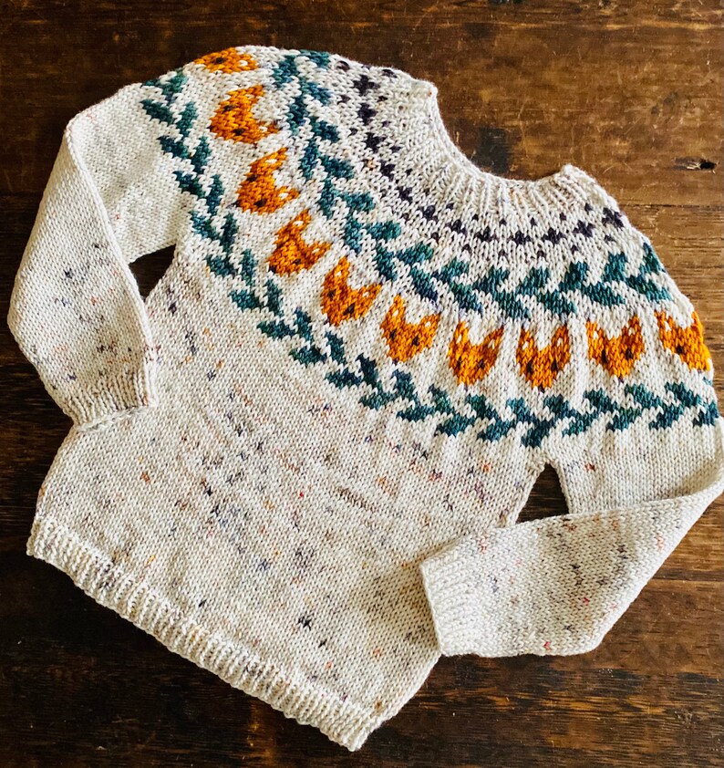 May include: A white knitted jumper with a colourful Fair Isle pattern featuring orange, green and blue accents. The jumper has a crew neck and long sleeves.