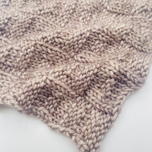Könnte beinhalten: Nahaufnahme eines beigefarbenen Strickstoffes mit einem strukturierten Muster. Der Stoff ist weich und hat einen dezenten Glanz.