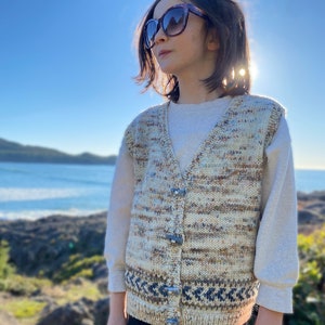 May include: A young person wearing a beige and brown knitted vest with a blue and white geometric pattern at the bottom. The vest has buttons down the front. The person is also wearing a white long-sleeved shirt, black pants, and sunglasses.
