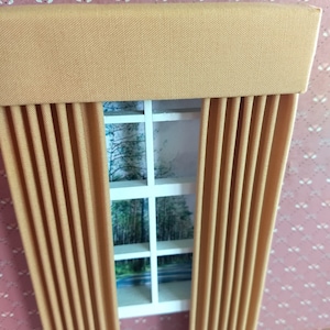 May include: Miniature window with tan curtains and a valance. The curtains are pleated and frame a view of a forest scene. The window frame is white, and the wall is pink with a floral pattern.