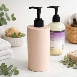 May include: A light pink ceramic soap dispenser with a black pump. The dispenser is cylindrical and sits on a white surface. A second bottle with a purple label is behind it. The scene includes a basket, soap, and greenery.