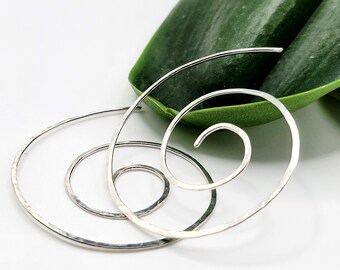 May include: Silver spiral earrings with a hammered texture. The earrings are arranged in a nested spiral design, with a smooth, polished finish. The earrings are set against a white background with a large green leaf.